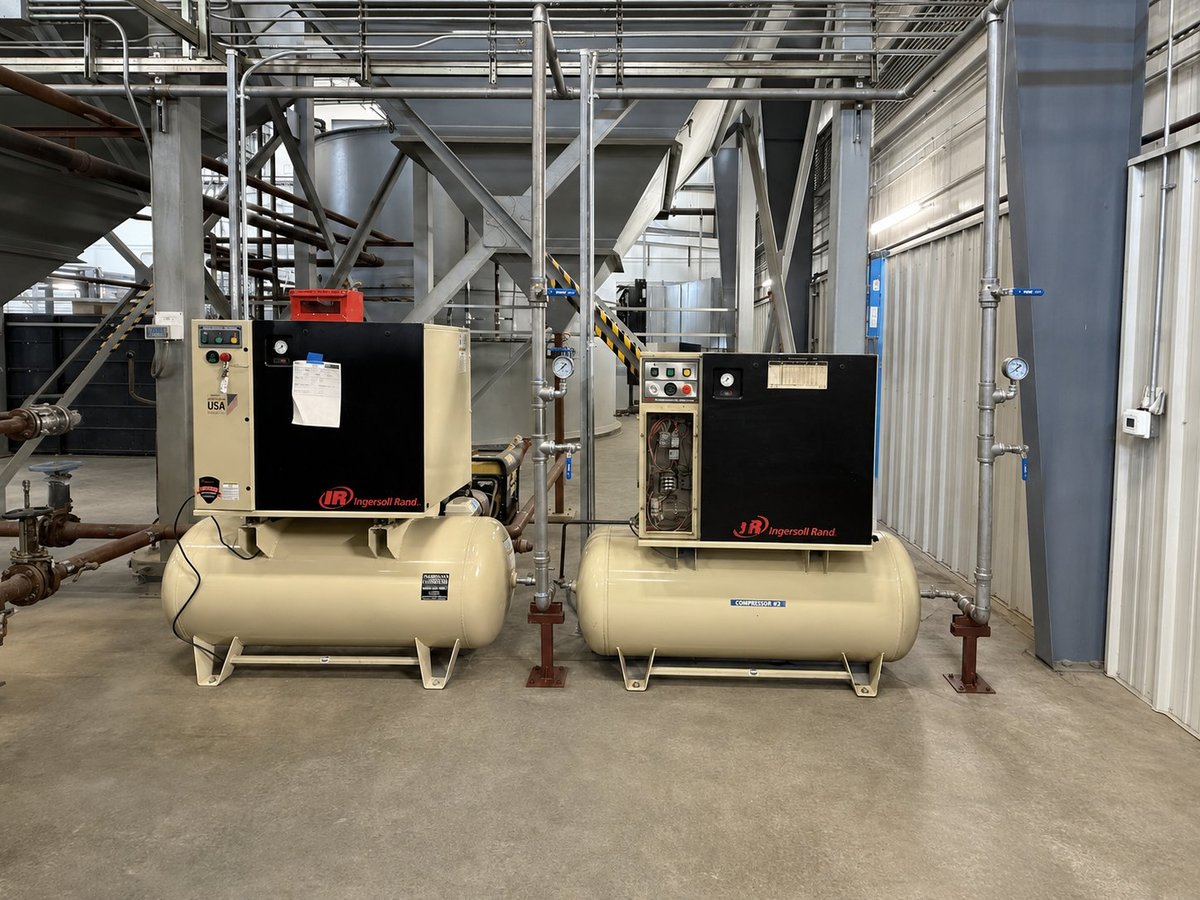 Two tan industrial air compressors with black motor units in a warehouse facility with metal framework and pipes overhead