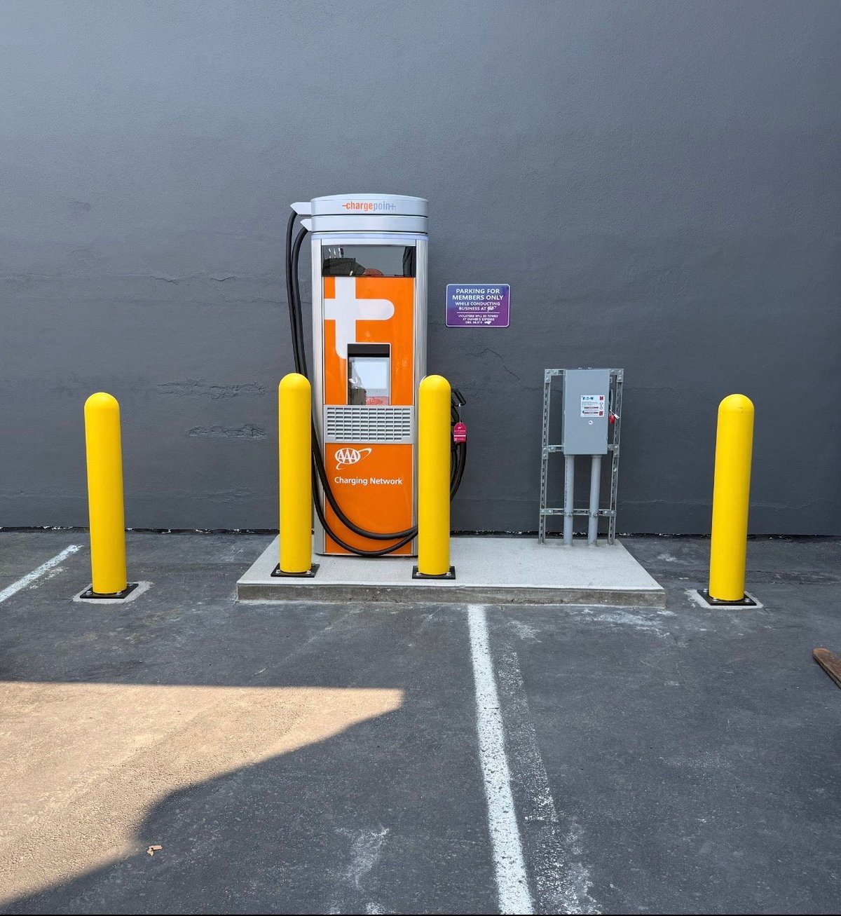 Electric vehicle charging station with orange branding, surrounded by four yellow protective bollards against a gray wall